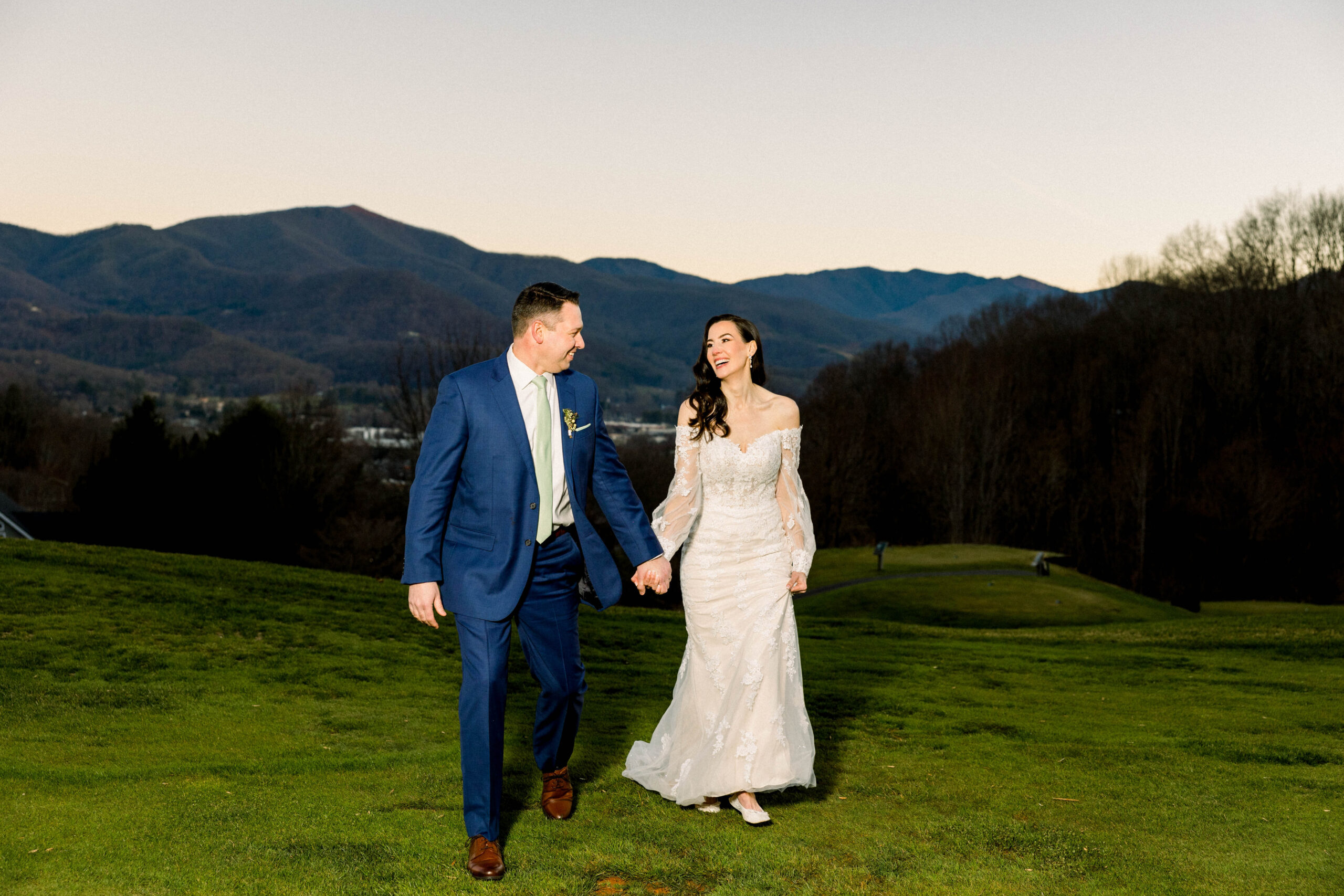 bride in white dress and groom in blue suit in front of mountains at sunset